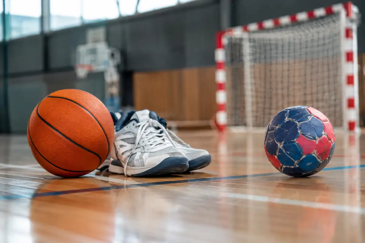 Ballon de basketball et ballon de handball posés côte à côte dans une salle de sport, près d’un panier et d’un but.