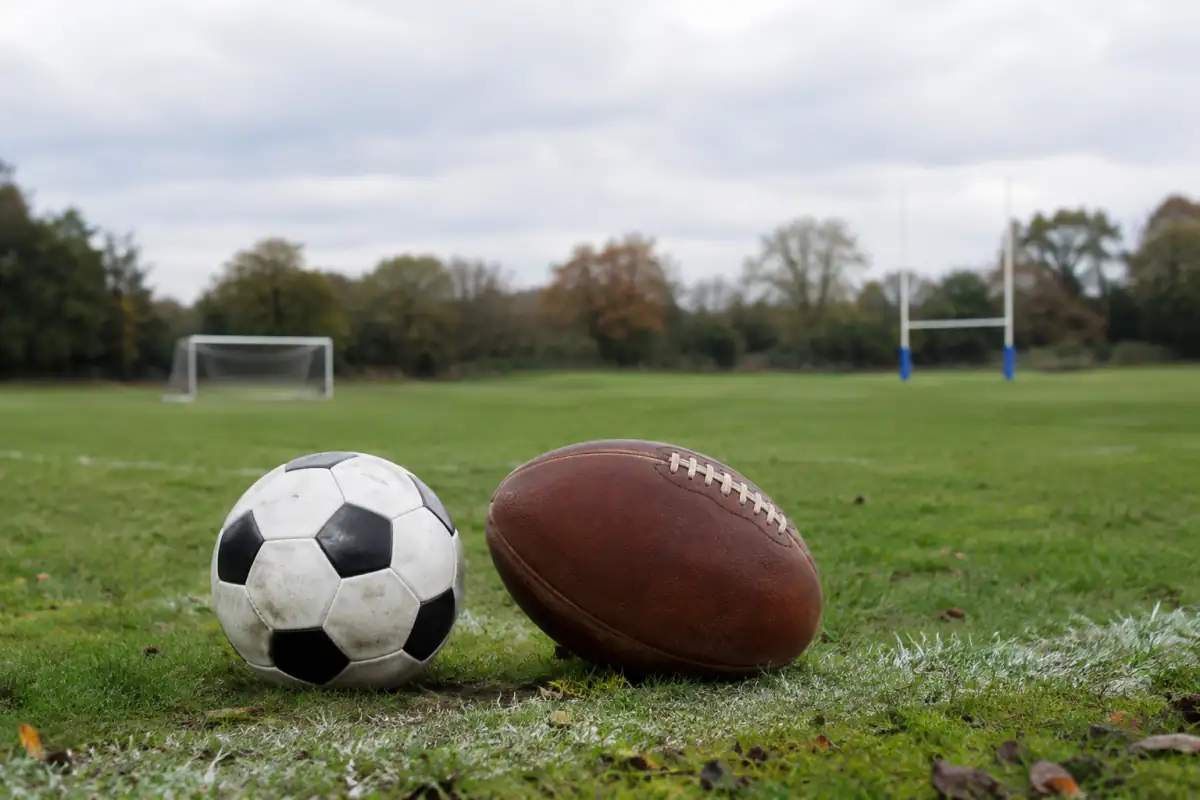 Ballon de football et ballon ovale côte à côte au bord d’un terrain, avec un but et des poteaux de rugby en arrière-plan.