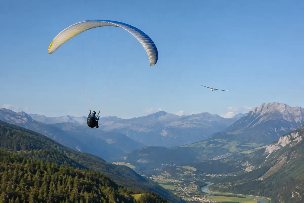 Un parapente et un planeur évoluent au-dessus d’une vallée française, illustrant la diversité des sports aériens.