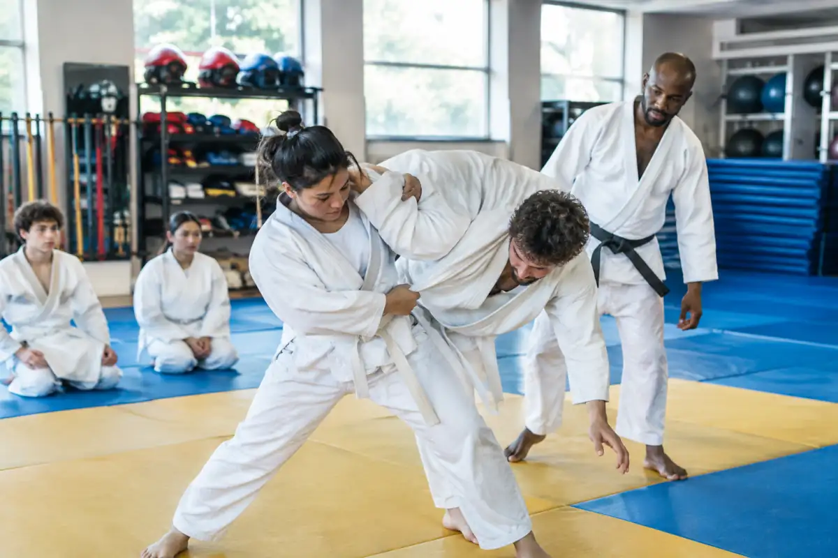 Deux adultes s’entraînent au judo sur un tatami dans un club, sous la supervision attentive d’un encadrant.