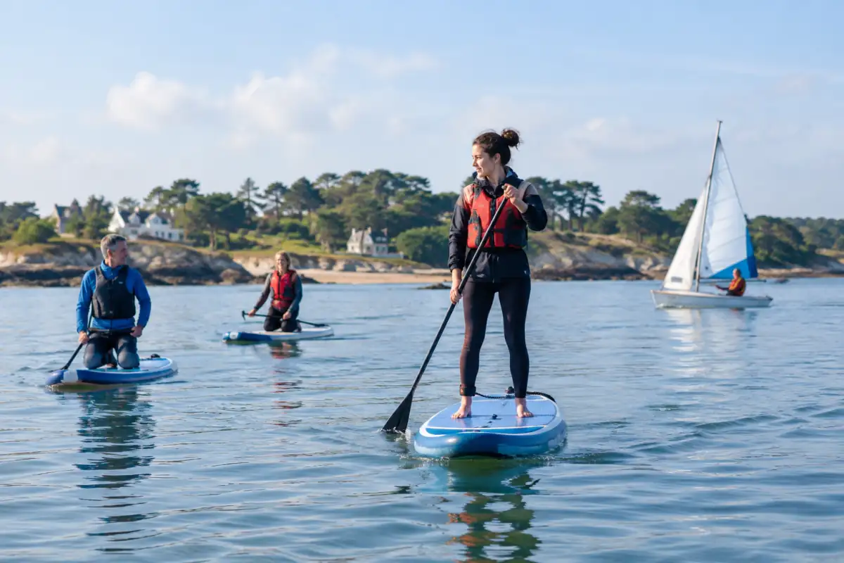 Des débutants en stand up paddle sur une baie calme, avec un petit voilier au loin, illustrent plusieurs sports nautiques.