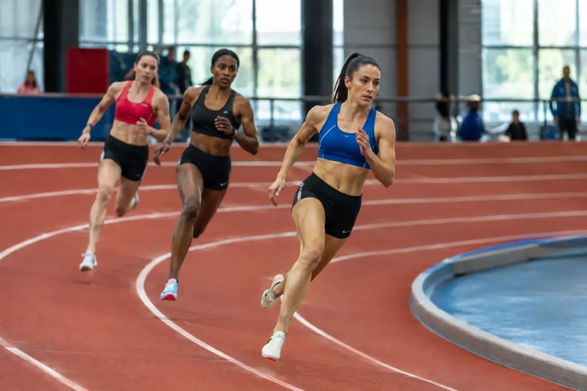 Athlètes en sprint dans le virage relevé d’une piste rouge lors d’une compétition d’athlétisme en salle.