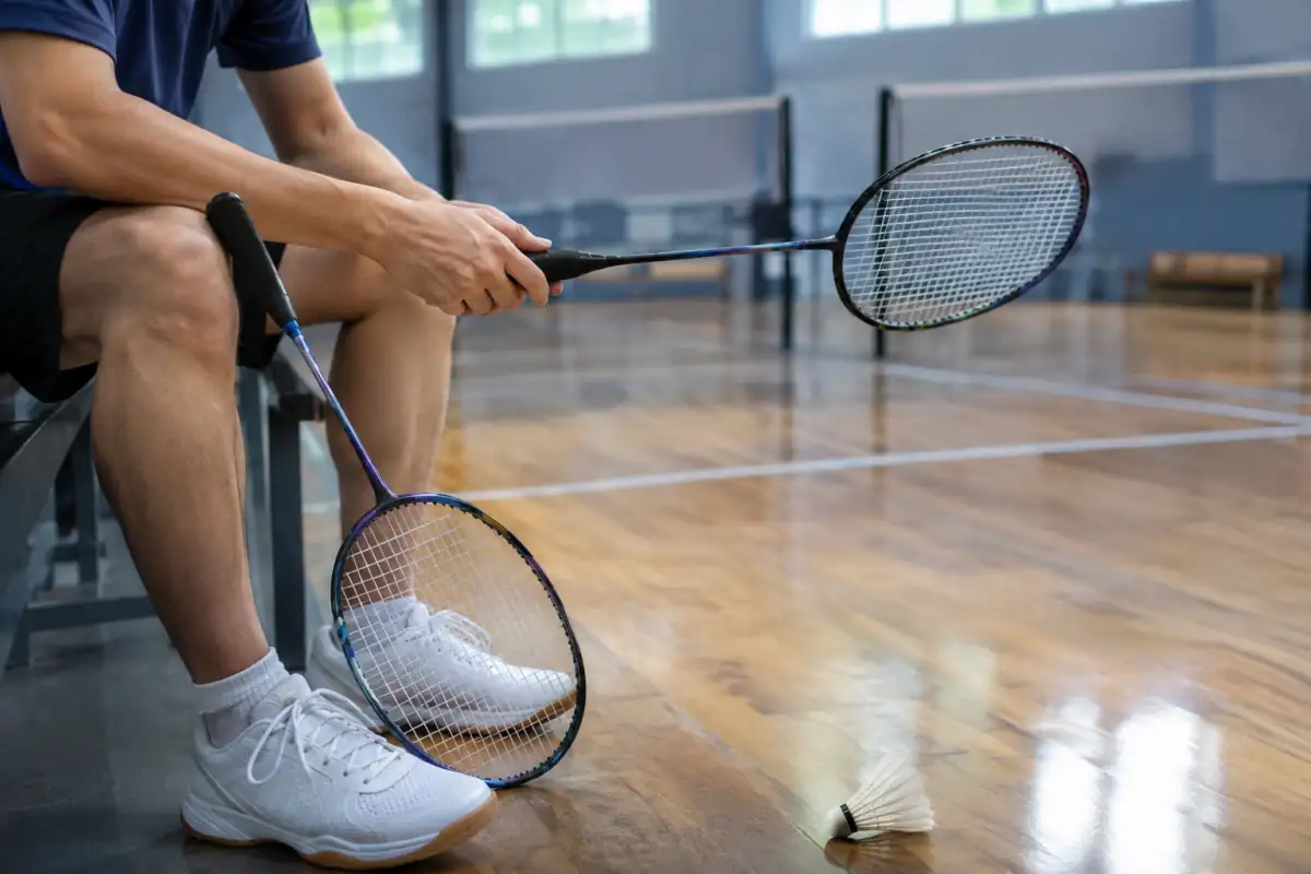 Une personne compare deux raquettes de badminton près de chaussures de salle et d’un volant en plumes, dans un gymnase.
