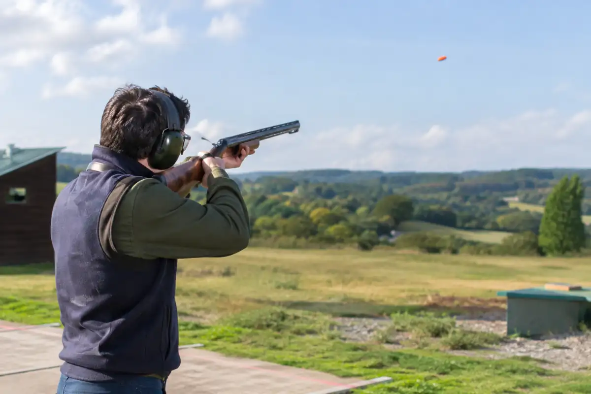 Un tireur équipé de lunettes et d’un casque suit un plateau lancé au-dessus d’un stand de ball-trap en plein air.
