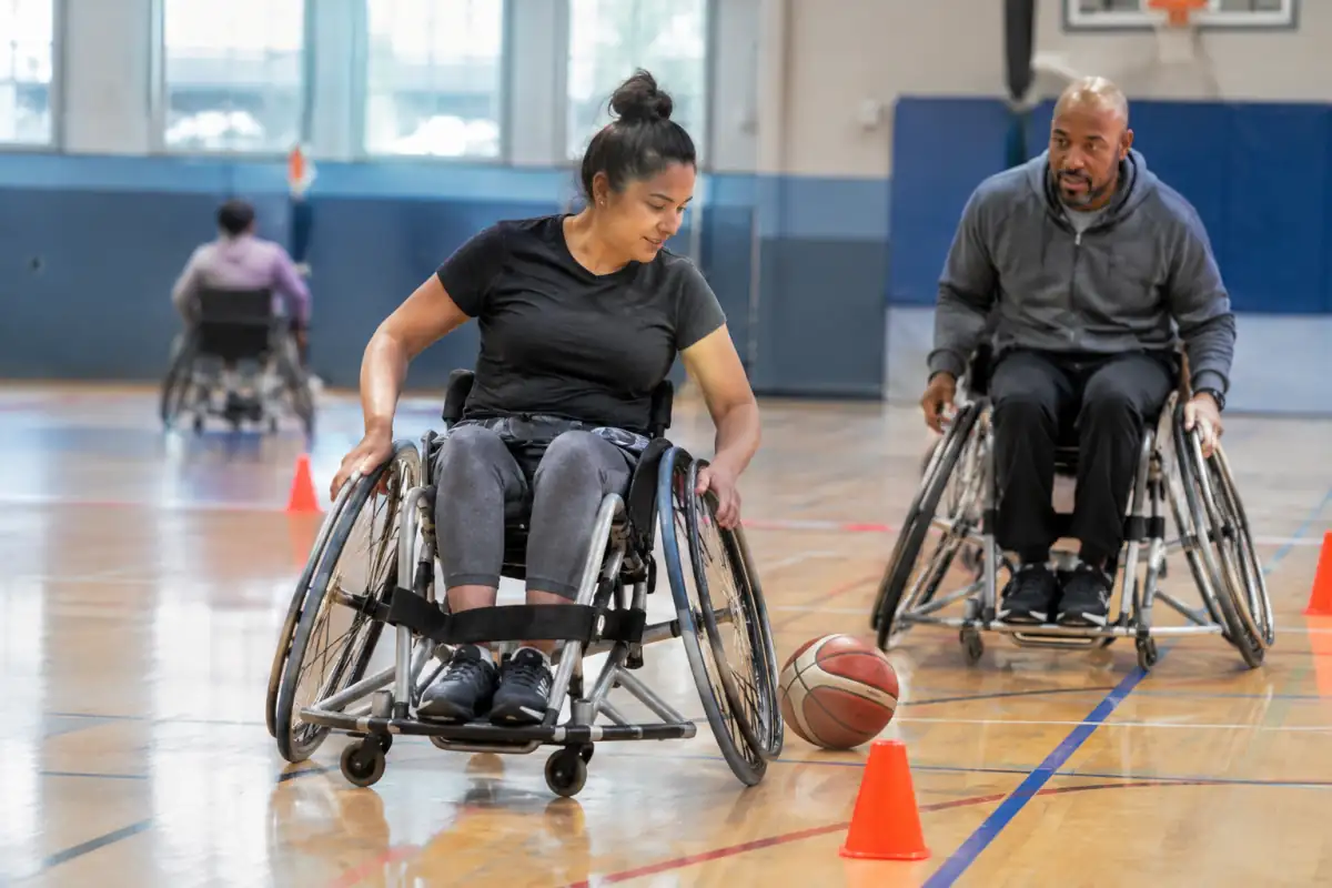 Un débutant en fauteuil de basket s’exerce à contourner des plots dans un gymnase, sous le regard attentif de son entraîneur.