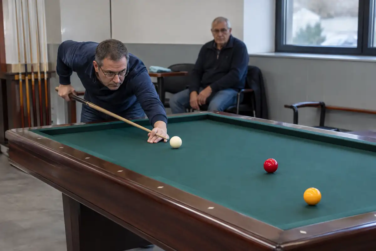Un joueur vise la bille blanche sur une table sans poches lors d’une partie de billard à trois bandes.