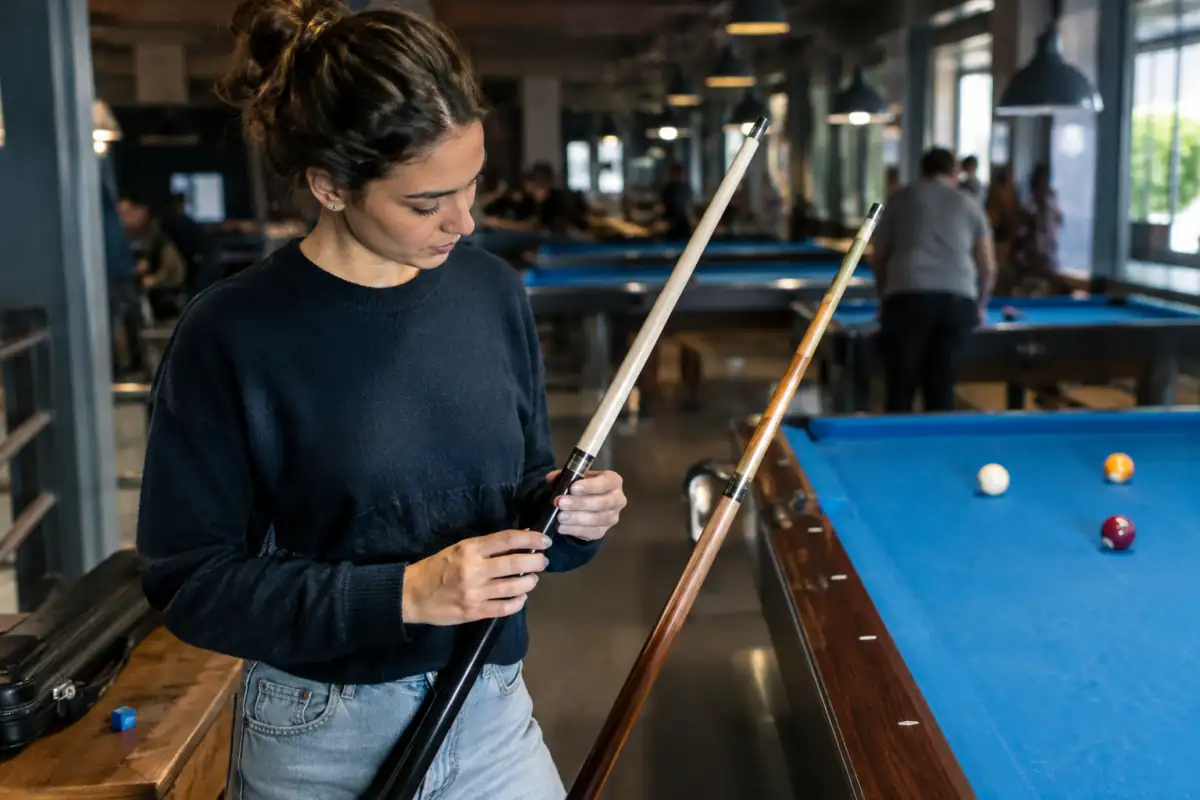 Un débutant compare deux queues de billard américain en bois près d’une table, avec un étui et de la craie.