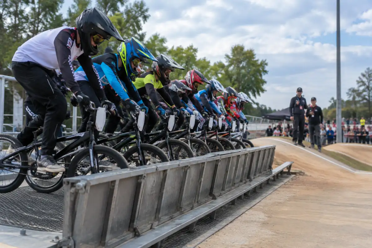 Huit pilotes de BMX attendent le signal derrière la grille de départ avant une manche qualificative sur une piste de course.