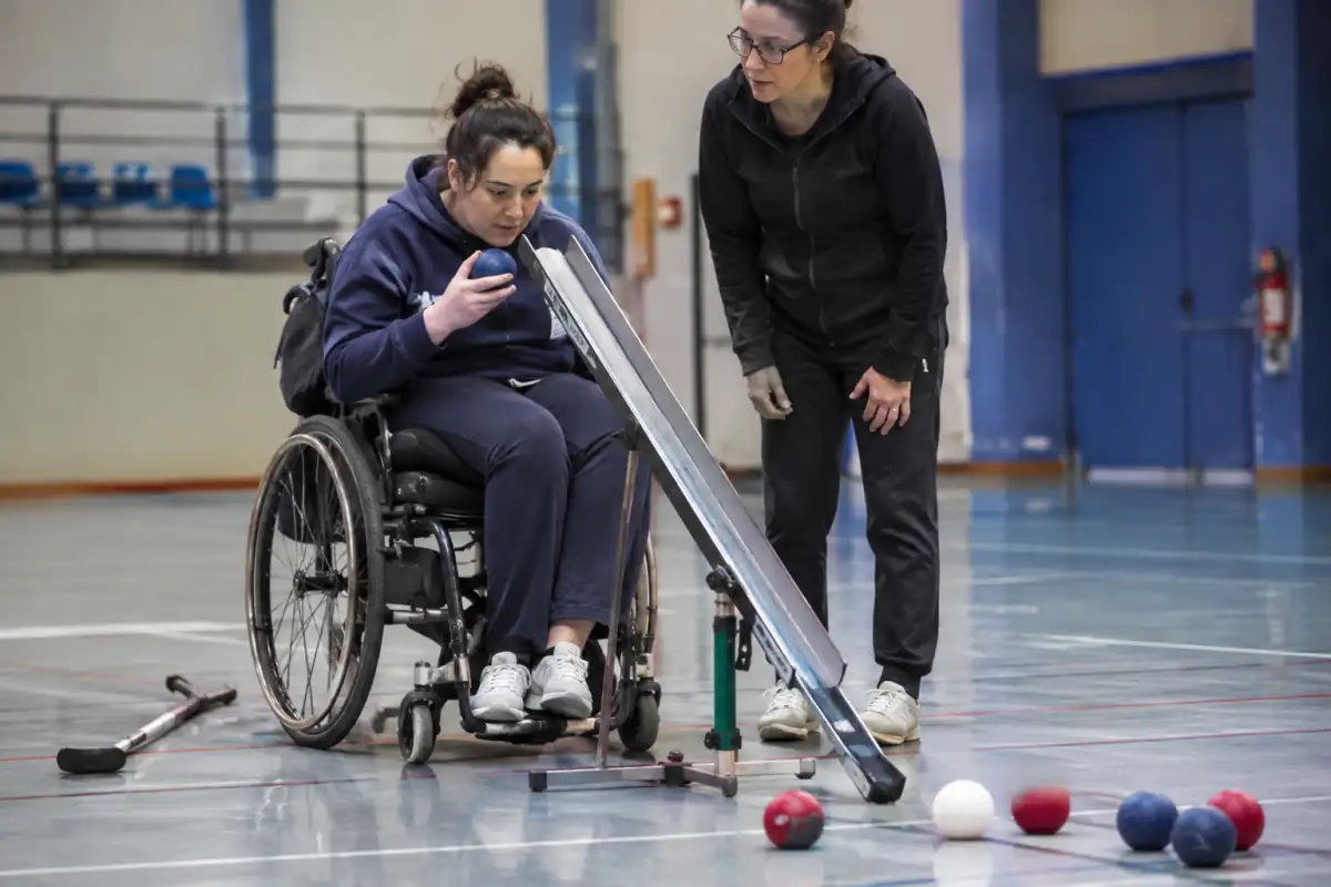 Joueur de boccia testant une balle près d’une rampe réglable, avec balles rouges, bleues et jack blanc sur le terrain.