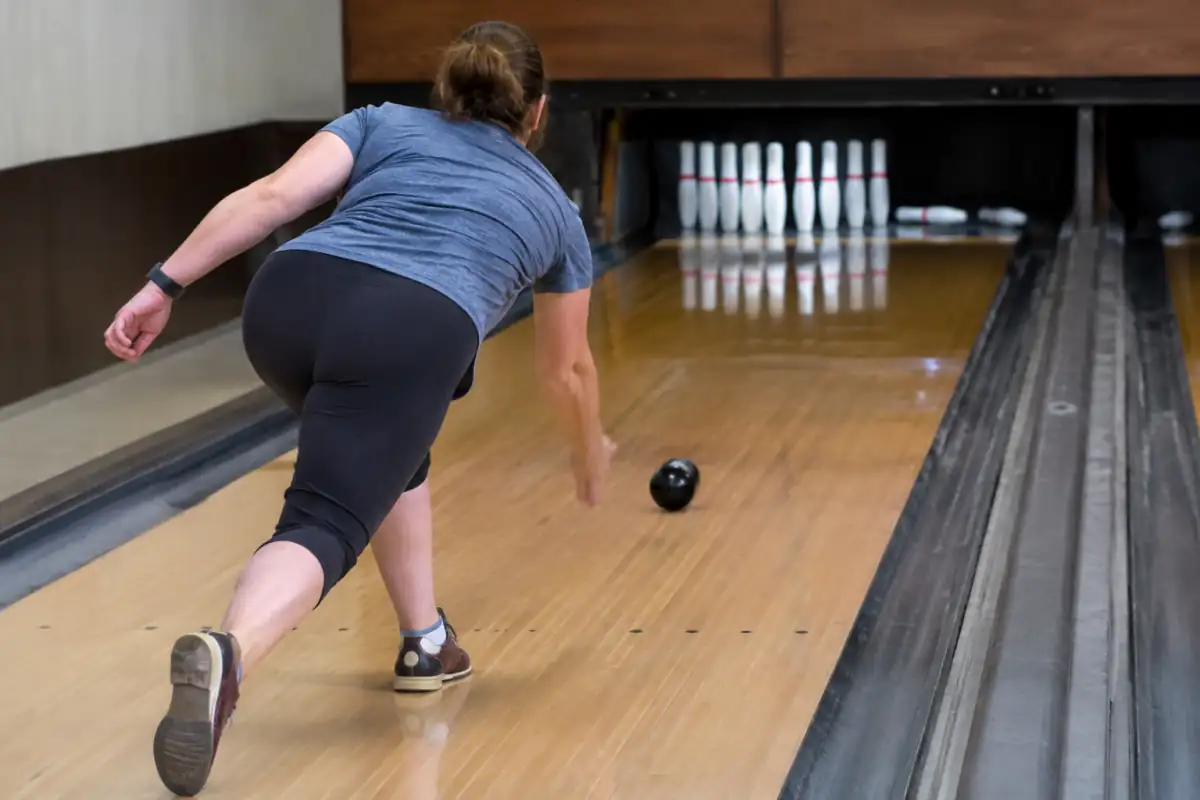 Une joueuse lance une petite boule sans trous vers les dix quilles fines sur une piste de candlepin bowling en salle.