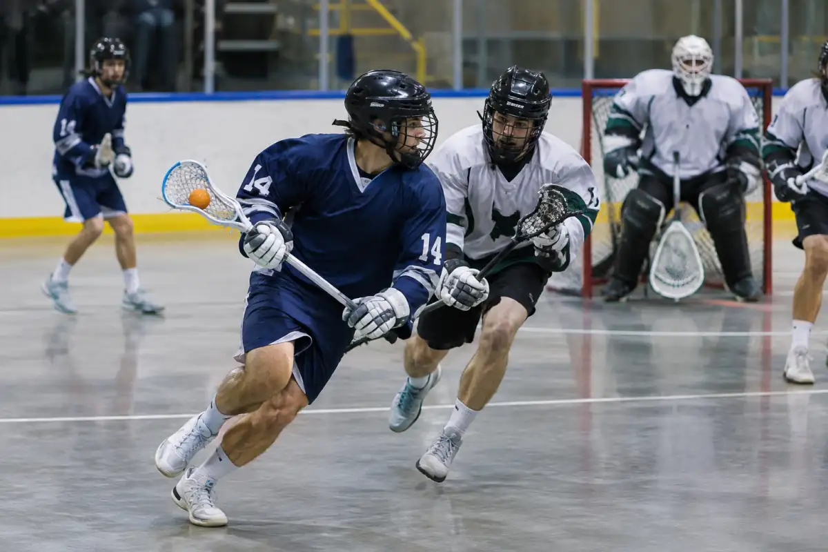Un joueur tire au but en box lacrosse dans une salle, tandis qu’un défenseur tente de le contrer devant le gardien protégé.
