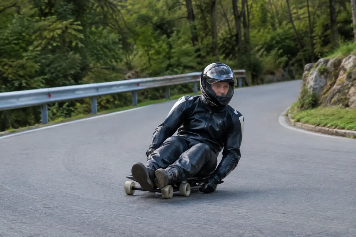 Un pilote casqué descend une route fermée, assis bas sur un buttboard à roulettes et portant une combinaison de protection.