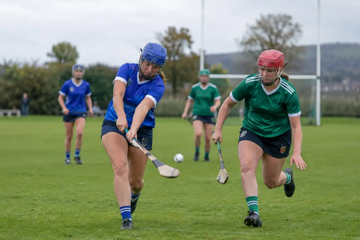 Joueuses de camogie en casques avec grille disputant la sliotar sur un terrain en herbe avec des crosses.