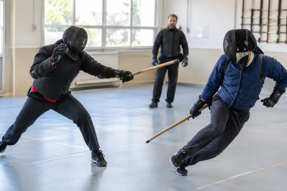 Deux pratiquants de canne de combat en assaut dans une salle, équipés de casques grillagés et de vestes de protection.