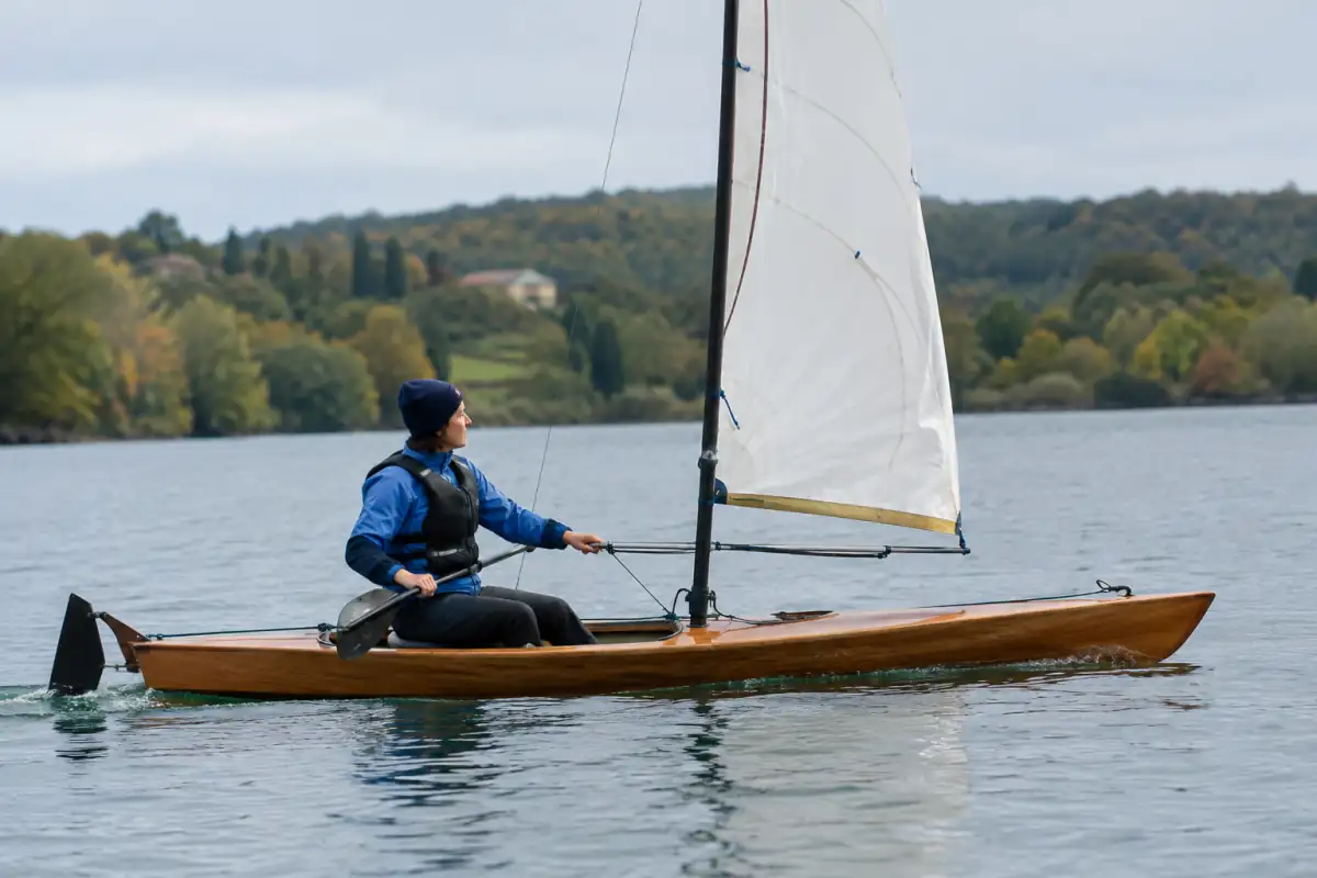 Un pratiquant règle la voile de son canoë à voile sur un lac, équipé d’un gilet et d’une pagaie.
