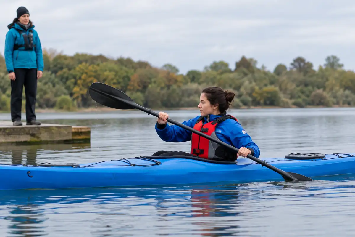 Un débutant travaille la rotation du buste en kayak sur une eau calme, sous l’observation d’un encadrant équipé.