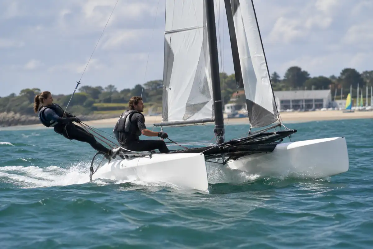 Deux équipiers en gilet et harnais équilibrent un catamaran de sport lancé sur l’eau près d’une côte française.