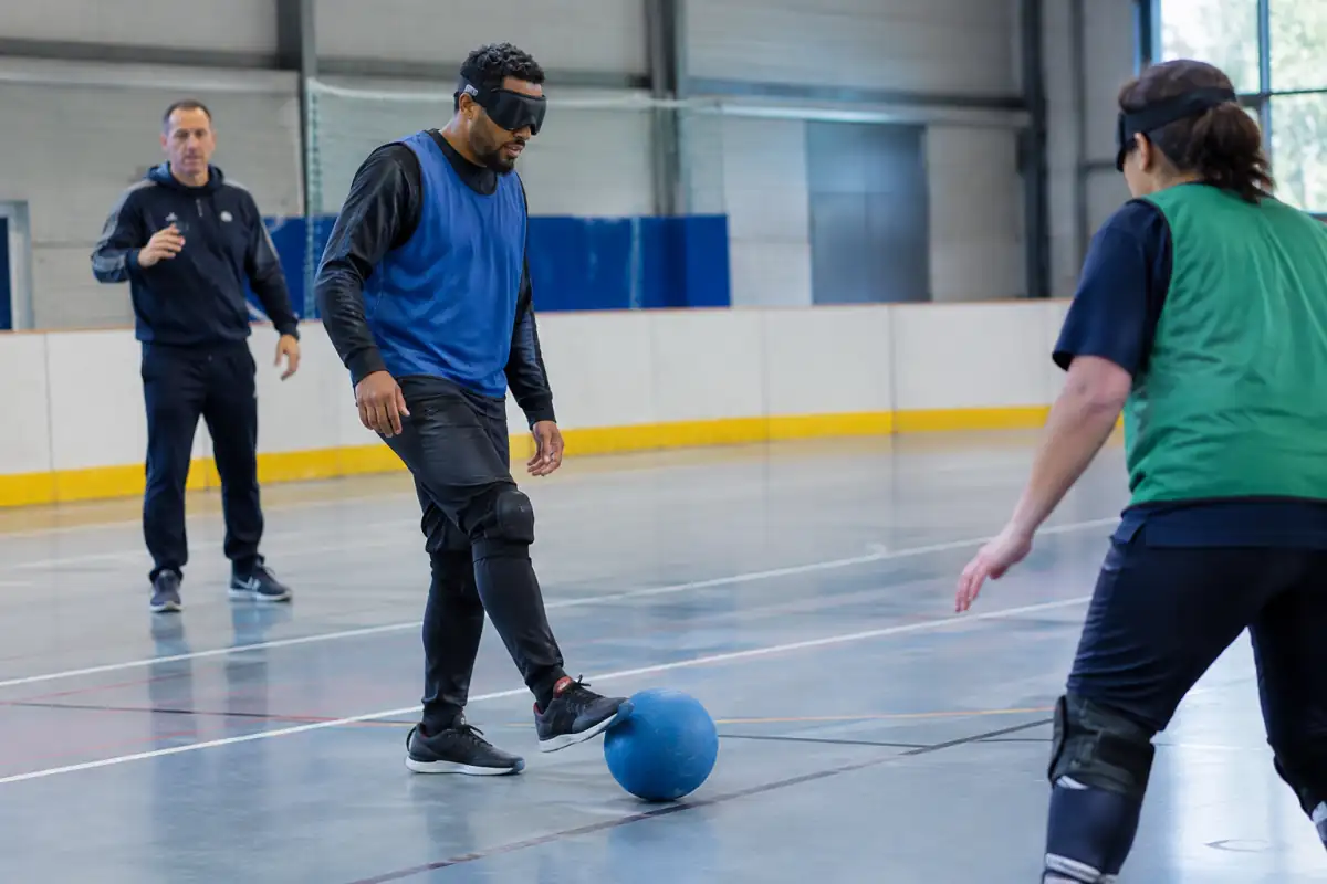 Un débutant en cécifoot écoute le ballon sonore avant de préparer une passe courte, guidé par un entraîneur dans un gymnase.