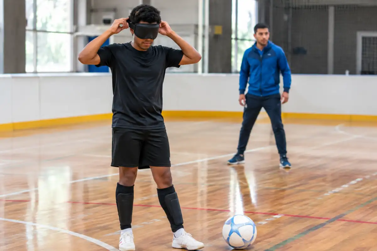 Joueur de cécifoot ajustant son masque occultant près d’un ballon sonore, avec chaussures de salle et protège-tibias.
