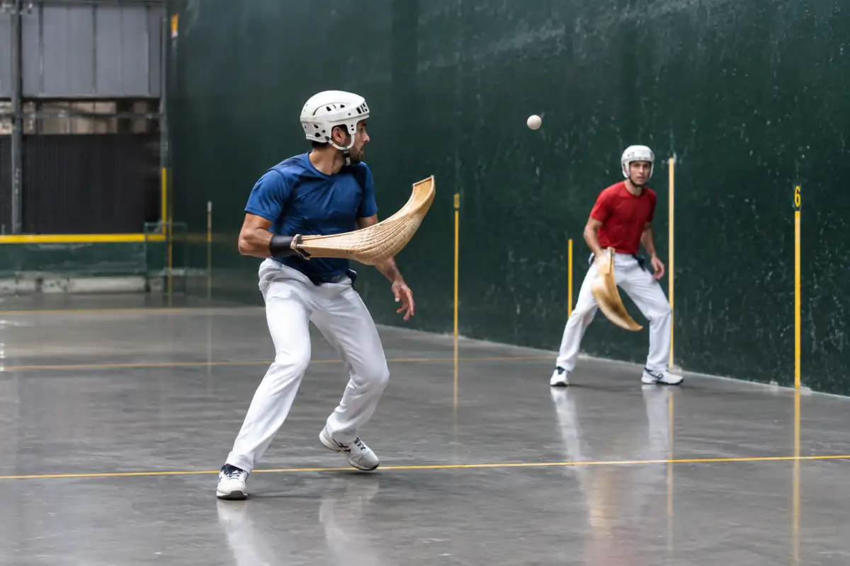 Un joueur de cesta punta réceptionne la pelote avec son panier incurvé dans un fronton couvert, face à son adversaire.