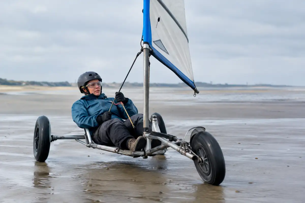 Un pilote casqué dirige un char à voile à trois roues sur une plage dégagée, voile tendue par le vent.