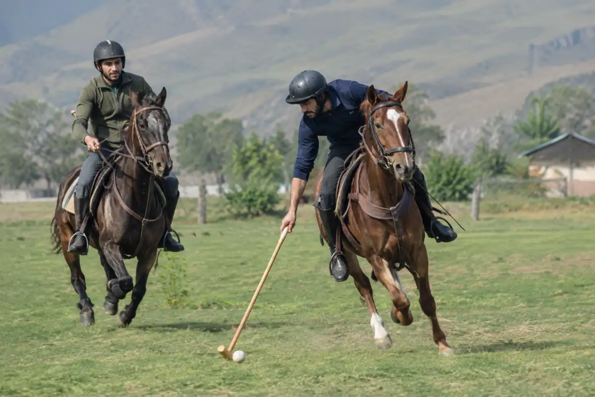 Cavaliers en casque jouant au chovgan sur un terrain extérieur, l’un frappant la balle au maillet depuis sa monture.