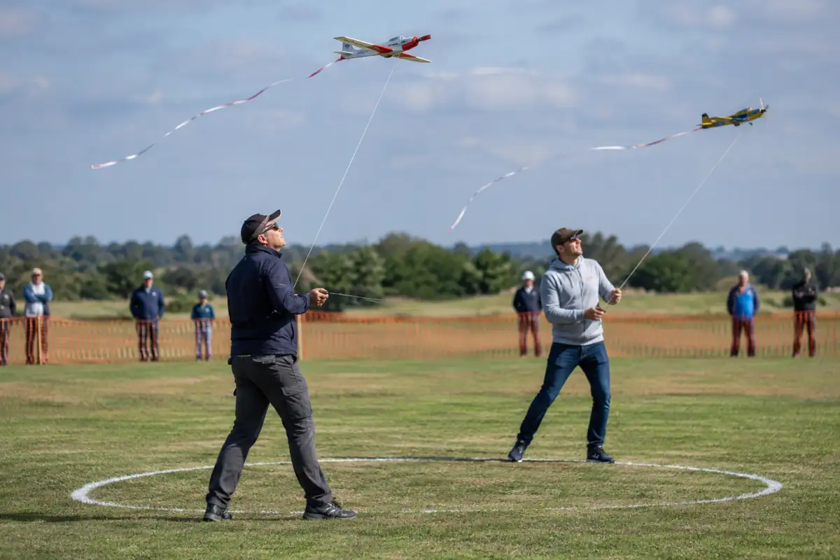 Deux pilotes manœuvrent leurs avions de combat aéromodélistes reliés par des lignes, rubans de traînée visibles au-dessus d’un terrain.