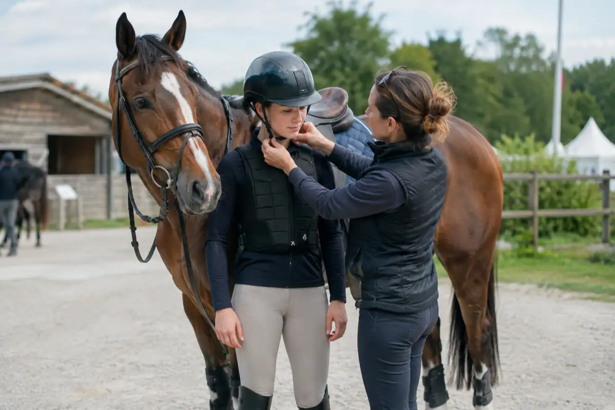 Une cavalière vérifie l’ajustement de son casque et de son gilet près d’un cheval équipé avant le cross.