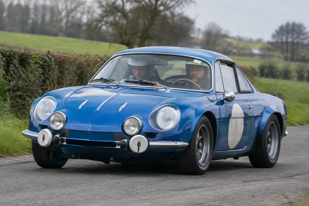 Équipage en voiture classique lors d’un rallye de régularité, le copilote suit un roadbook sur une route de campagne.
