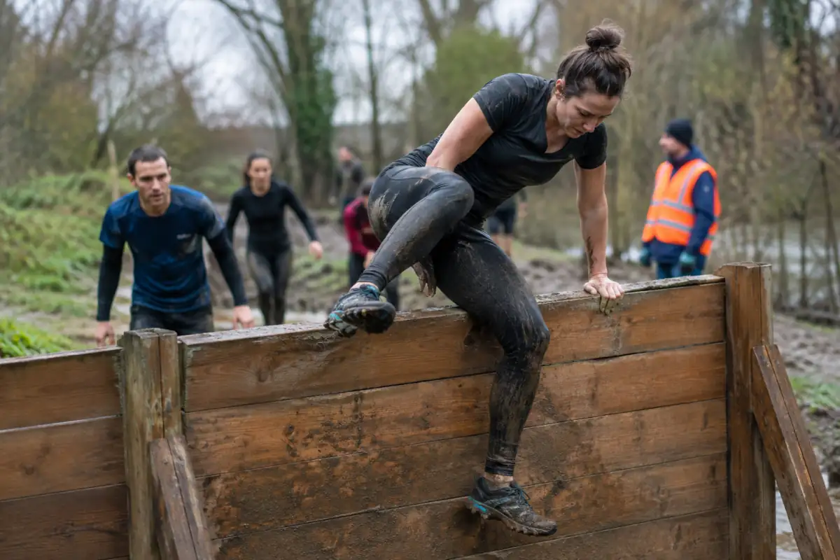 Une participante franchit un mur en bois sur un parcours boueux de course d’obstacles en pleine nature.