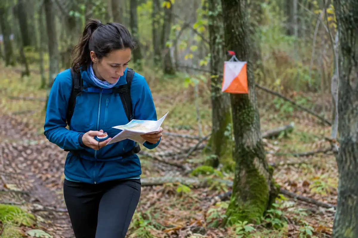 Une orienteuse consulte sa carte et sa boussole en forêt avant de rejoindre une balise de course d’orientation.