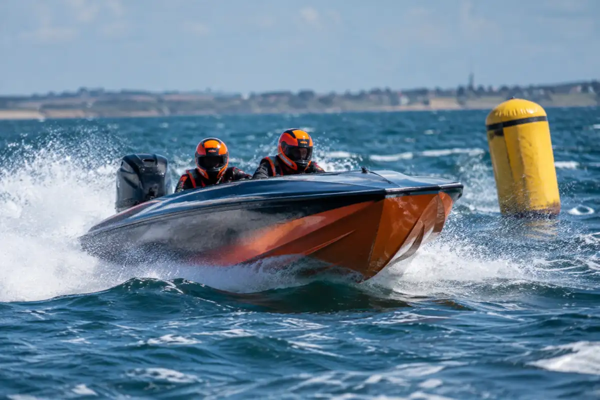 Équipage casqué dans un bateau de course offshore lancé sur une boucle balisée en mer près du littoral.