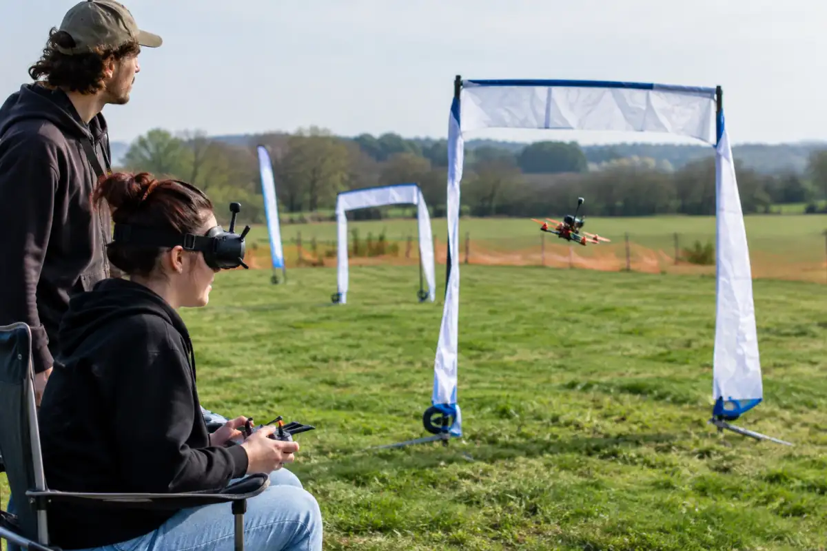 Pilote en lunettes FPV et observateur suivant un drone de course qui franchit une porte sur un circuit de club en France.
