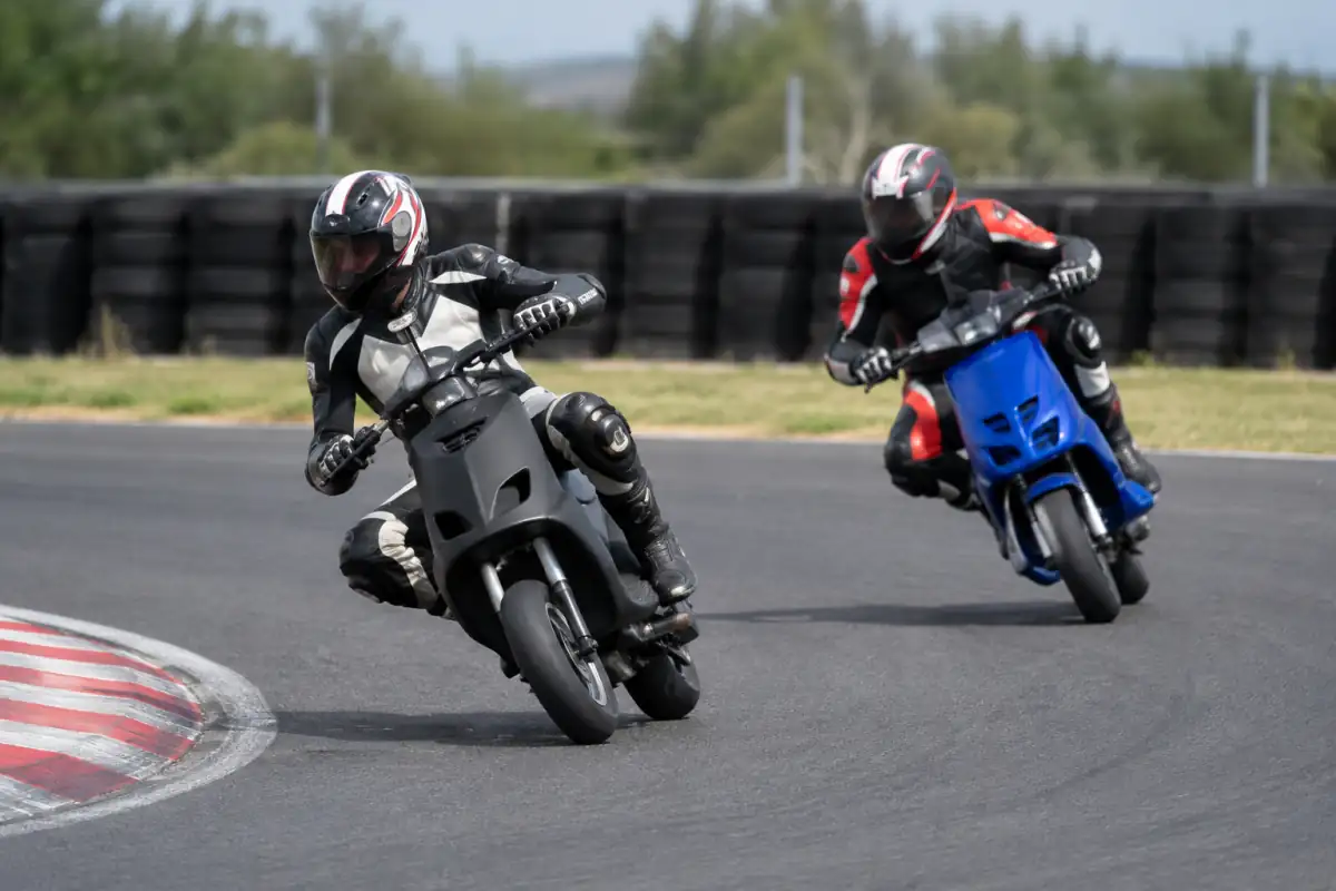 Deux pilotes en combinaison et casque négocient un virage sur leurs scooters de course, sur un circuit fermé.