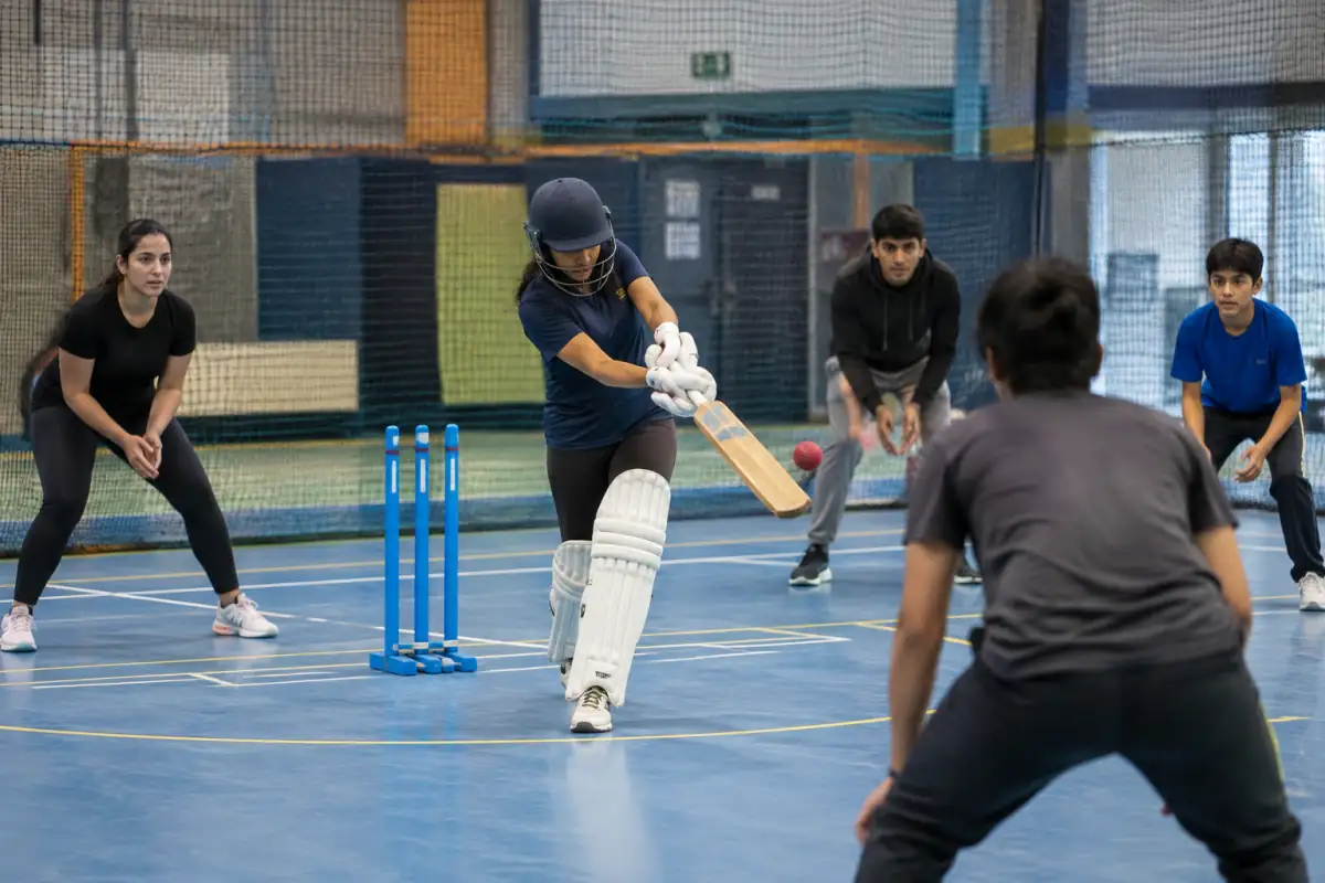 Un batteur équipé joue un coup de batte face aux défenseurs dans un terrain de cricket en salle bordé de filets.