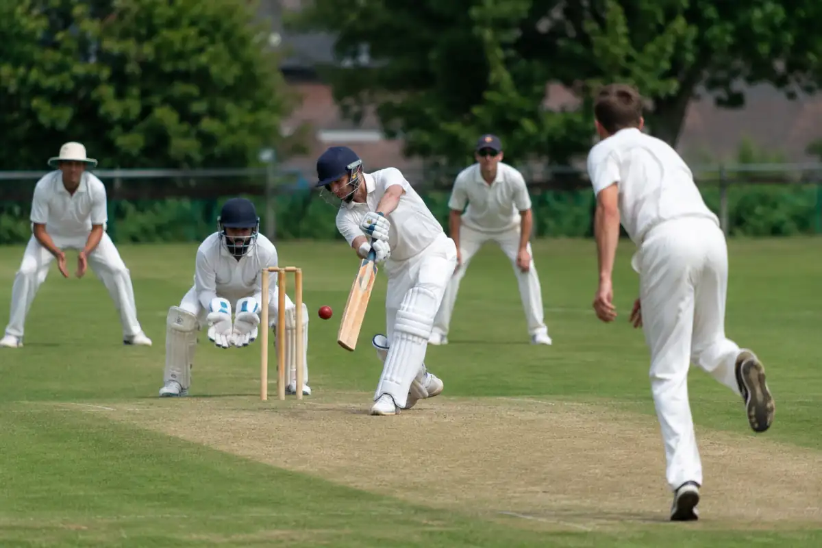 Un batteur équipé frappe la balle sur le pitch pendant un match de cricket en plein air, avec le gardien derrière le guichet.