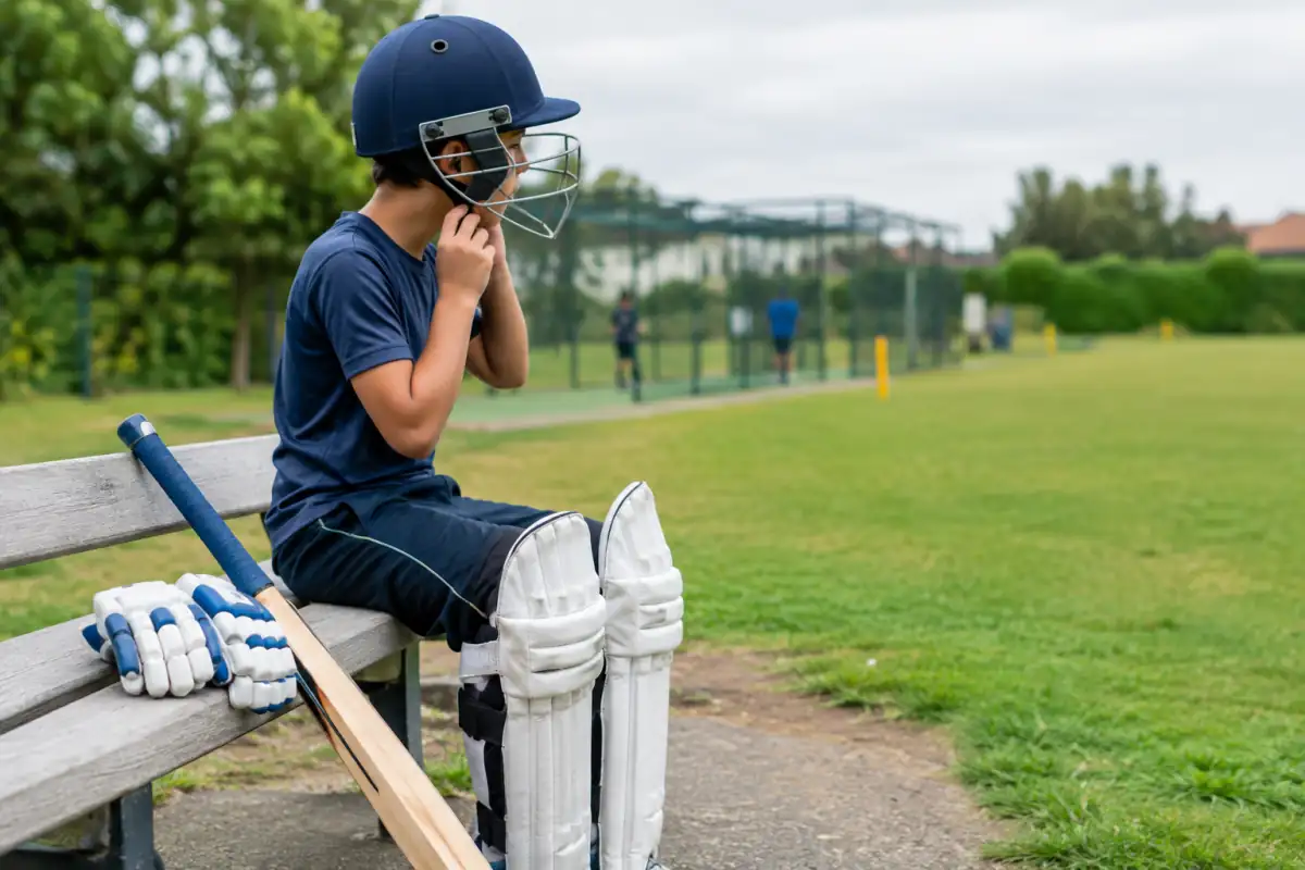 Un débutant ajuste un casque de cricket à grille près d’une batte, de gants et de jambières avant une séance.