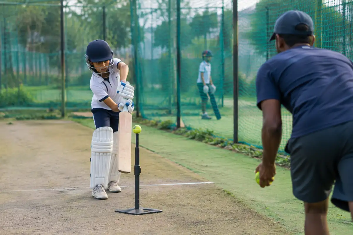 Un débutant en tenue de cricket travaille une frappe droite sur balle posée dans un filet d’entraînement, sous le regard d’un partenaire.