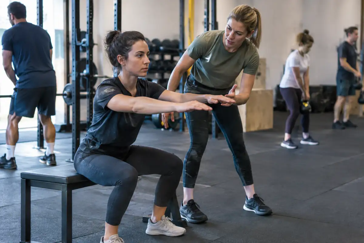 Un coach accompagne une débutante dans un squat vers un banc, lors d’une première séance de CrossFit adaptée.