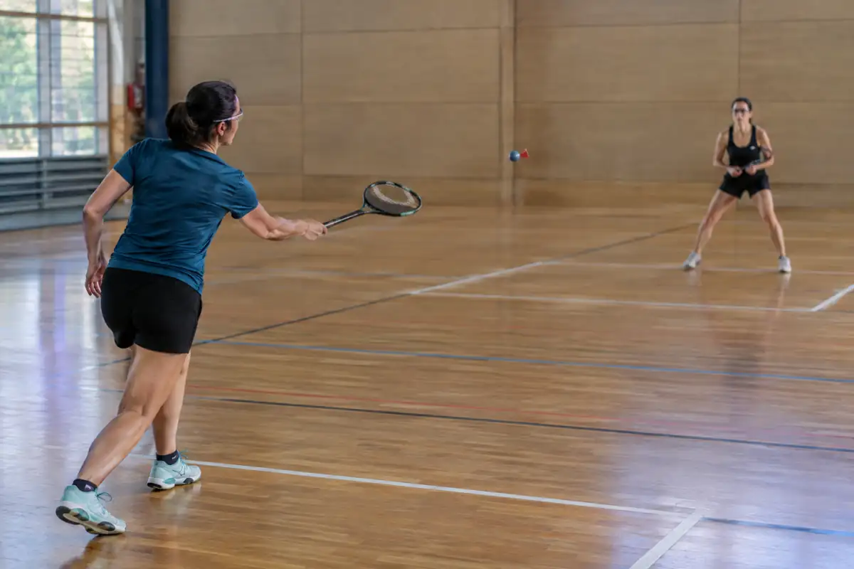 Deux joueurs équipés de raquettes et de lunettes disputent un échange de crossminton sans filet dans un gymnase.