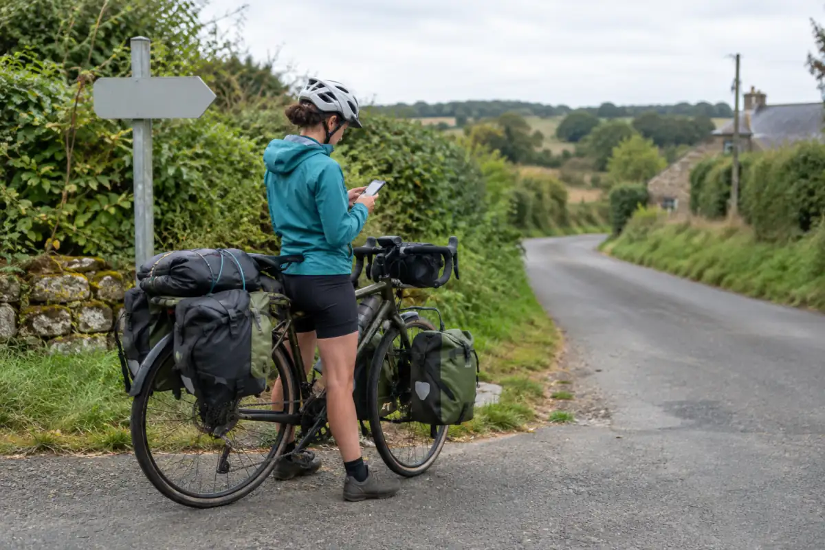 Cycliste en voyage à l’arrêt sur une petite route, consultant une carte hors ligne avec ses sacoches équilibrées avant de reprendre son itinéraire.