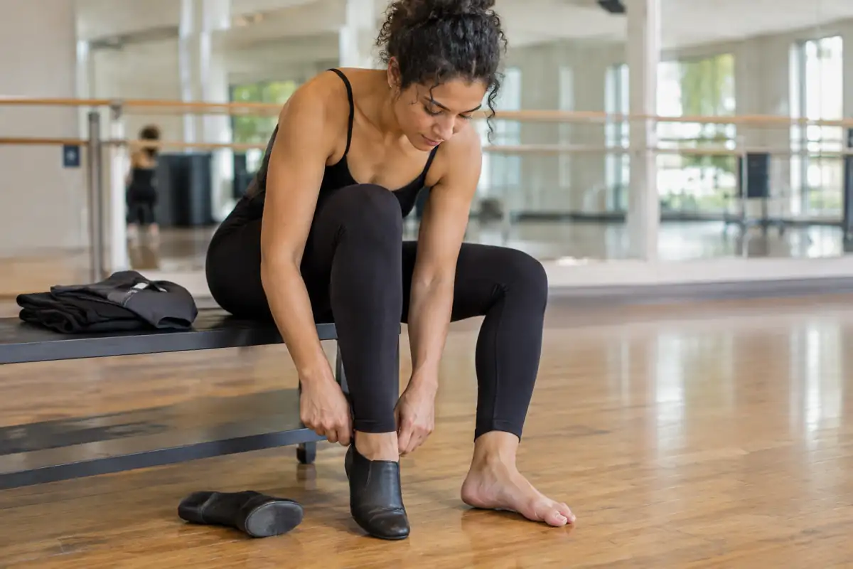 Danseuse débutante essayant un chausson de jazz dans un studio, avec sa tenue de cours pliée à côté sur un banc.