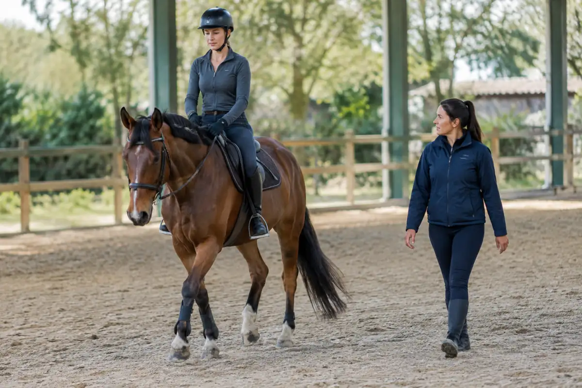 Un adulte débutant travaille les transitions au pas dans une carrière, accompagné par un moniteur et monté sur un cheval calme.