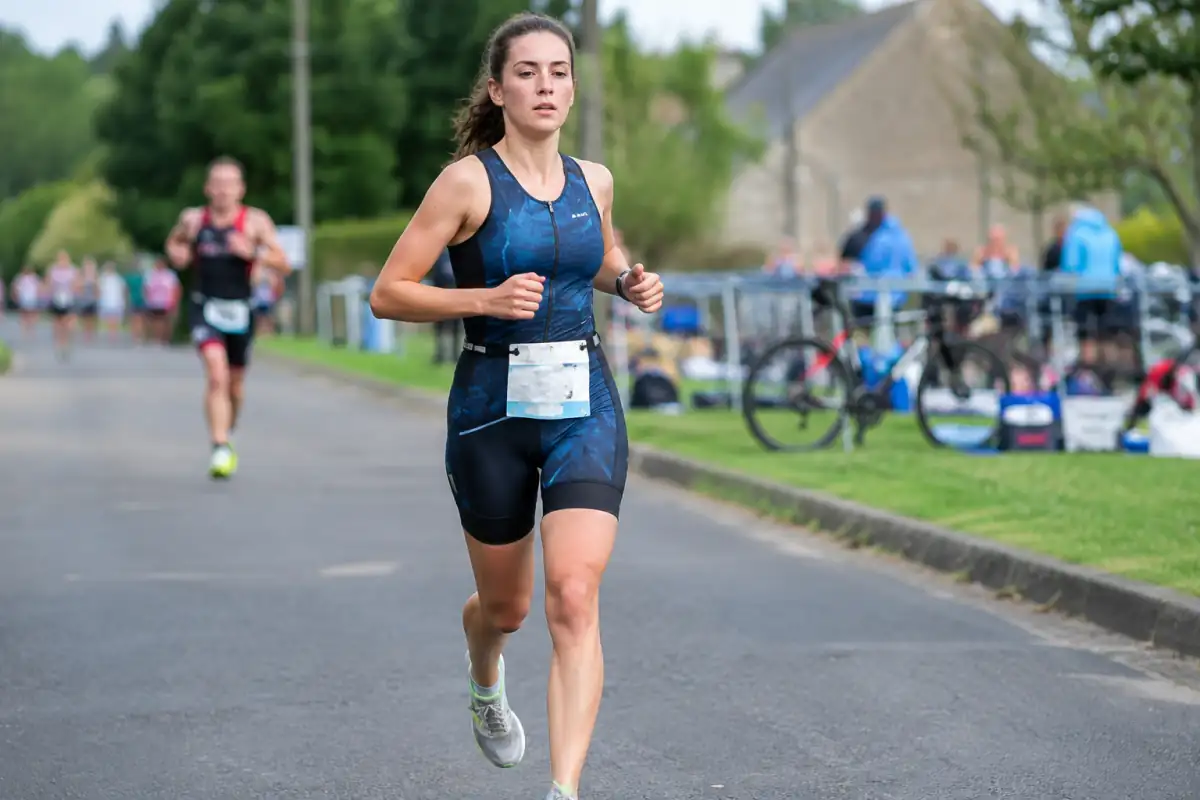 Athlète en course à pied lors de la dernière étape d’un duathlon, avec une zone de transition et des vélos en arrière-plan.