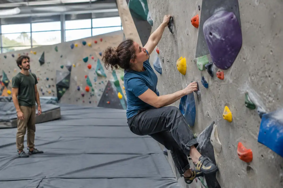 Grimpeur en chaussons réalise un mouvement d’escalade de bloc au-dessus des tapis de réception d’une salle.