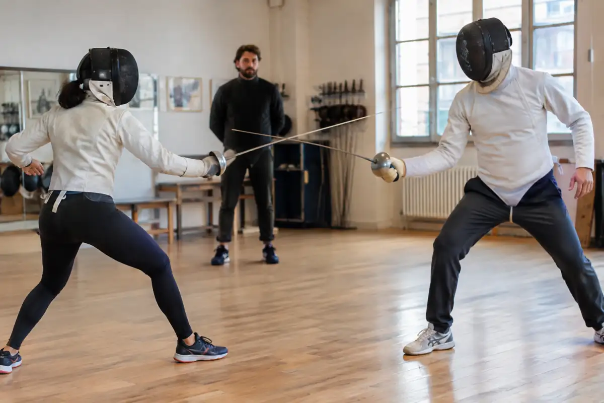 Deux escrimeurs masqués répètent un duel chorégraphié à la rapière émoussée dans une salle, sous le regard d’un encadrant.