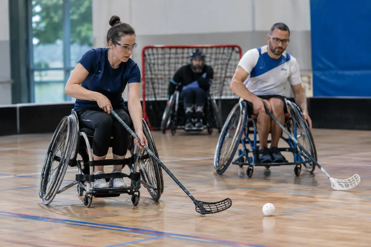 Deux joueurs en fauteuil roulant manient leur crosse et se disputent une balle perforée lors d’une partie de floorball fauteuil en salle.