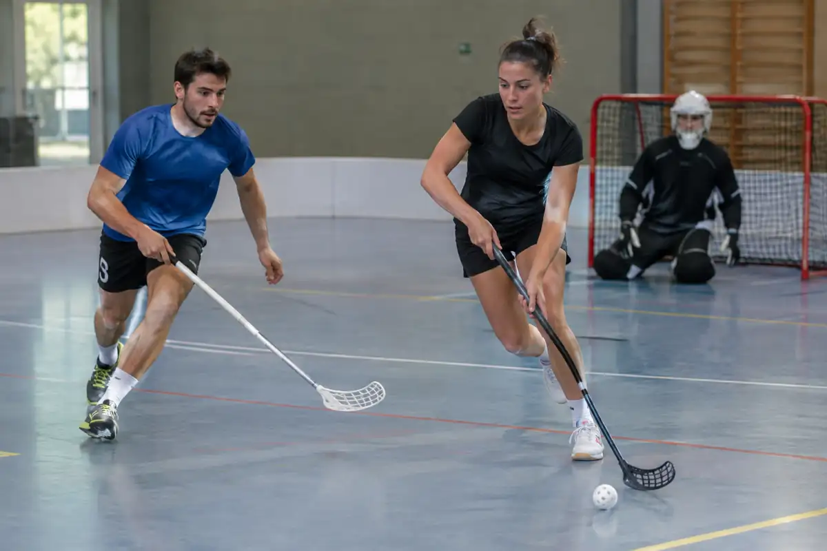 Match de floorball en salle : deux joueurs manient leur crosse près d’une balle perforée devant un gardien masqué.