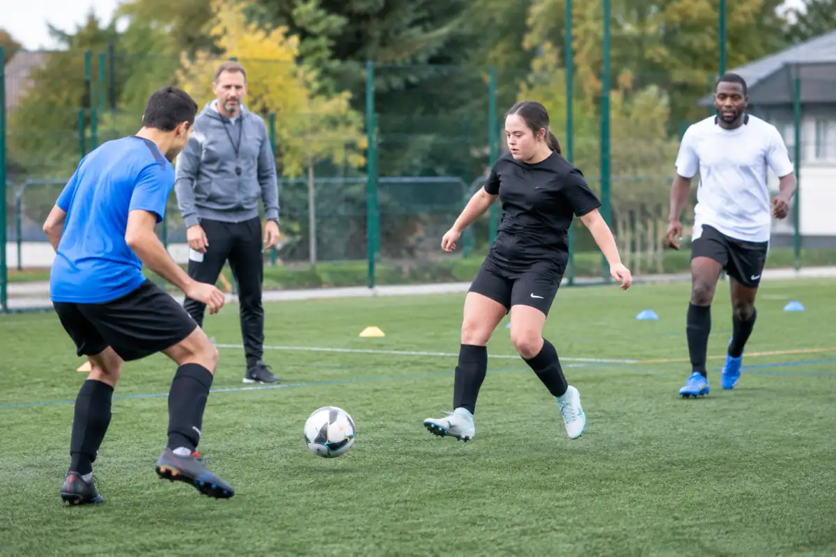 Des joueurs pratiquent le football adapté sur un terrain synthétique, en travaillant une passe sous le regard d’un éducateur.
