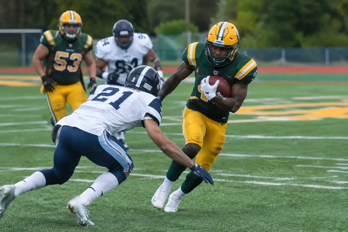 Action de football canadien sur grand terrain : un porteur du ballon tente d’avancer face à un défenseur équipé.